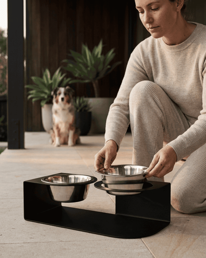 Woman setting up a black elevated pet feeder with two bowls on a wooden deck.