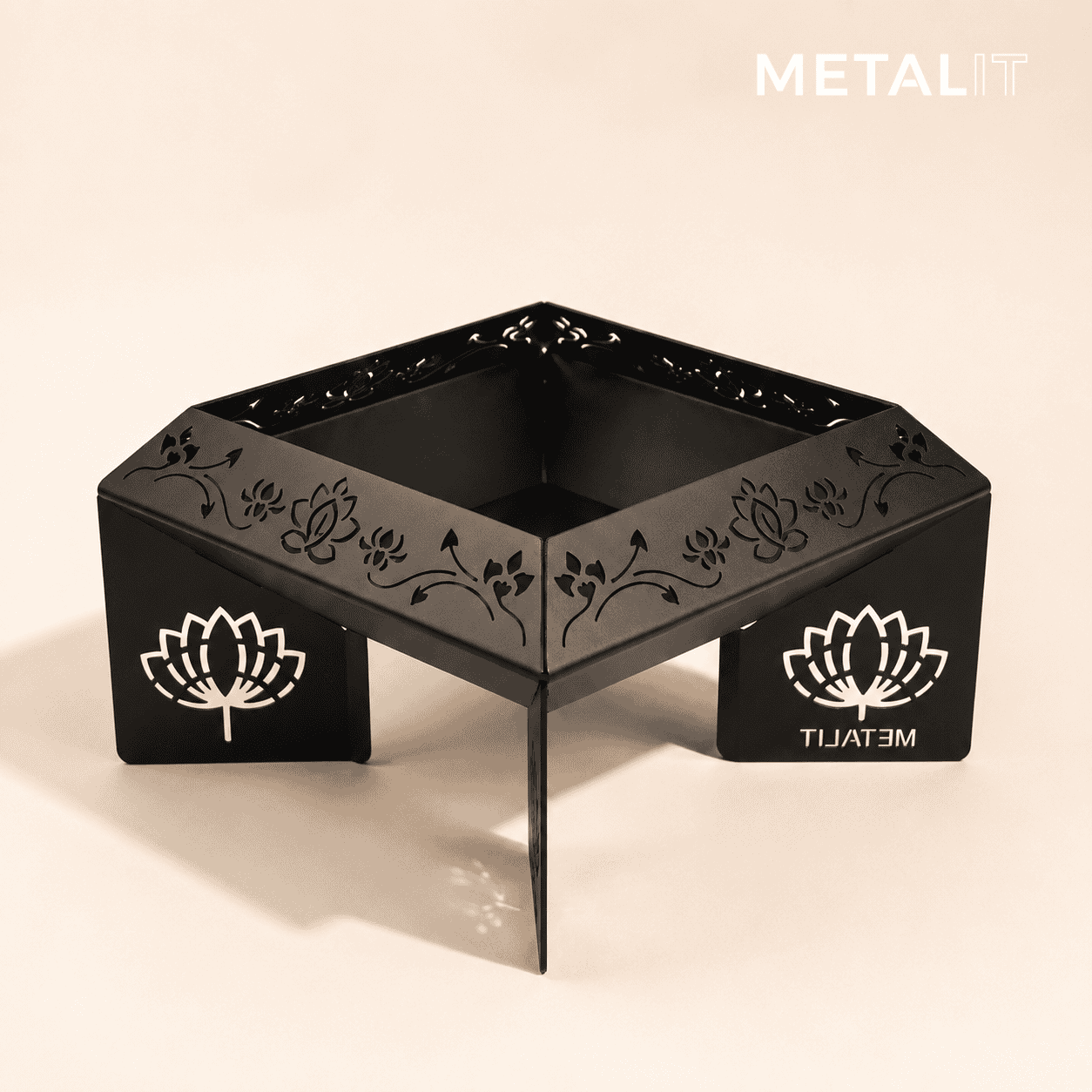 A black metal square havan kund with laser-engraved floral patterns on the sides and white lotus logo cutouts on the front panels, shown on a light beige background with a 'METALIT' brand watermark in the top right.